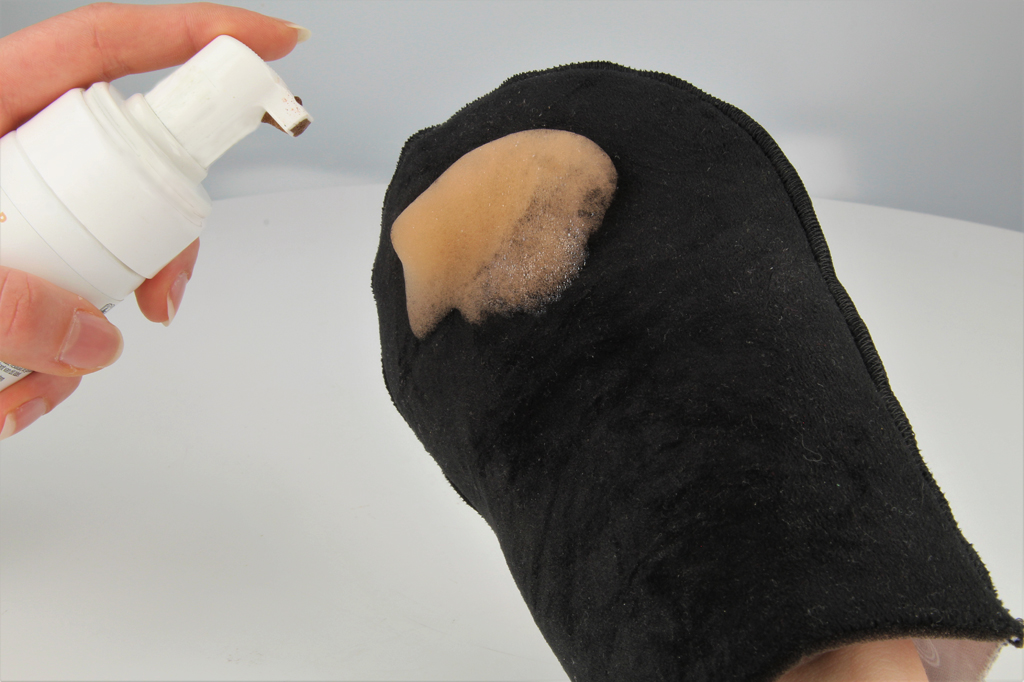 Zu sehen ist ein schwarzer Handschuh, auf dem aus einer weißen Tube ein brauner Schaum aufgetragen wurde, um diesen mit dem Handschuh auf der Haut zu verteilen.
